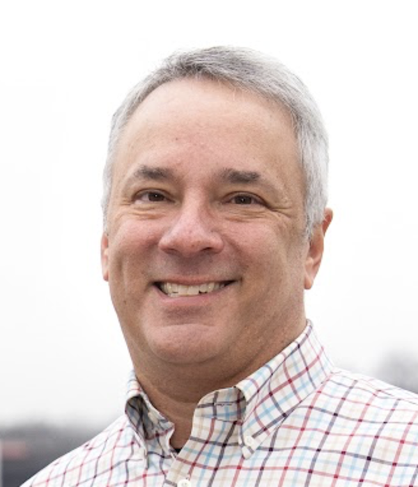 Charles Green smiling wearing a white shirt with red and blue checks with a white sky in the background.