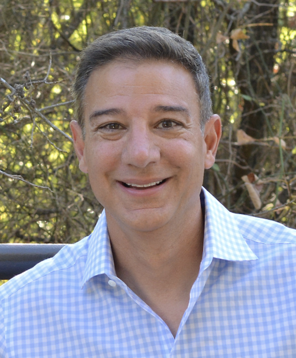 Ray Mallouk smiling in a white and light blue checked shirt with greenery in the background.