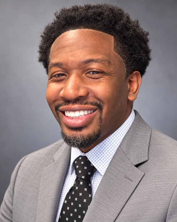 Greg Jackson in a tan suite and black tie with white spots and white dress shirt with a full smile.